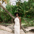 A woman with an afro stands on a sandy beach wearing a sleeveless, high-neck, floor-length white dress. Palm trees and a fallen tree trunk are visible in the tropical outdoor setting.