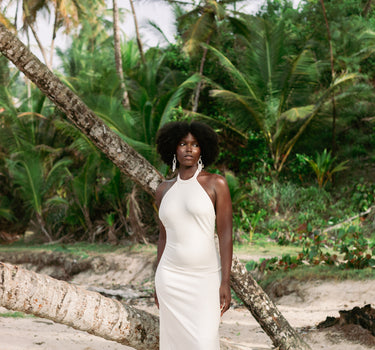 A woman with an afro stands on a sandy beach wearing a sleeveless, high-neck, floor-length white dress. Palm trees and a fallen tree trunk are visible in the tropical outdoor setting.