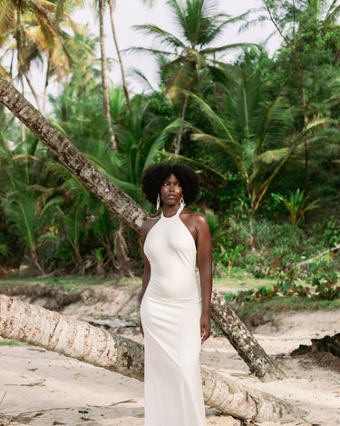 A woman with an afro stands on a sandy beach wearing a sleeveless, high-neck, floor-length white dress. Palm trees and a fallen tree trunk are visible in the tropical outdoor setting.