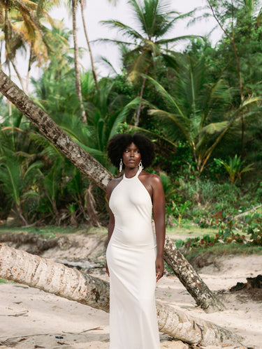 A woman with an afro stands on a sandy beach wearing a sleeveless, high-neck, floor-length white dress. Palm trees and a fallen tree trunk are visible in the tropical outdoor setting.