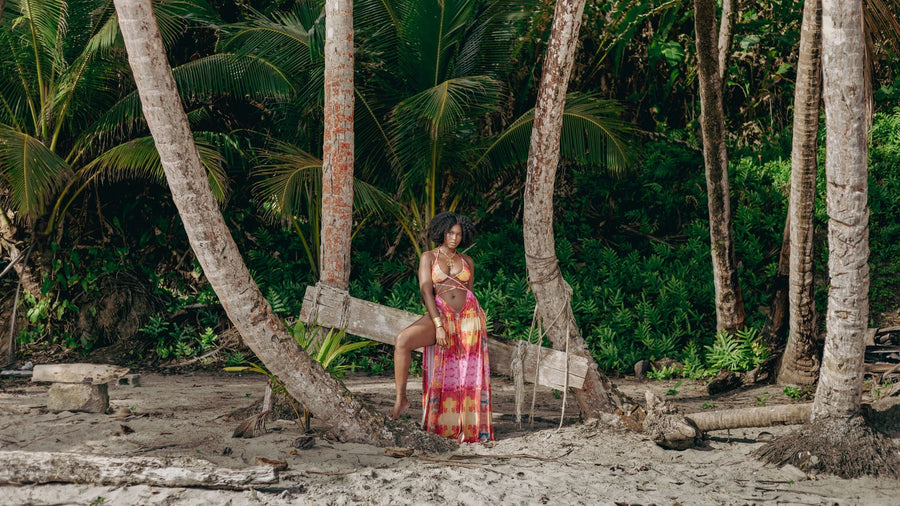A woman wearing a vibrant pink, orange, and yellow patterned maxi dress sits on a wooden platform on a tropical beach, surrounded by palm trees and sandy ground.